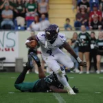 Ein Footballspieler in einem weißen Vikings-Trikot mit der Nummer 4 läuft mit dem Ball und weicht einem Tackle eines Dragons-Spielers in Grün aus, der auf dem Rasen liegt. Cheerleader und eine Menschenmenge schauen im Stadionhintergrund zu. Diese Beschreibung wurde mit der FootballR KI automatisch generiert.