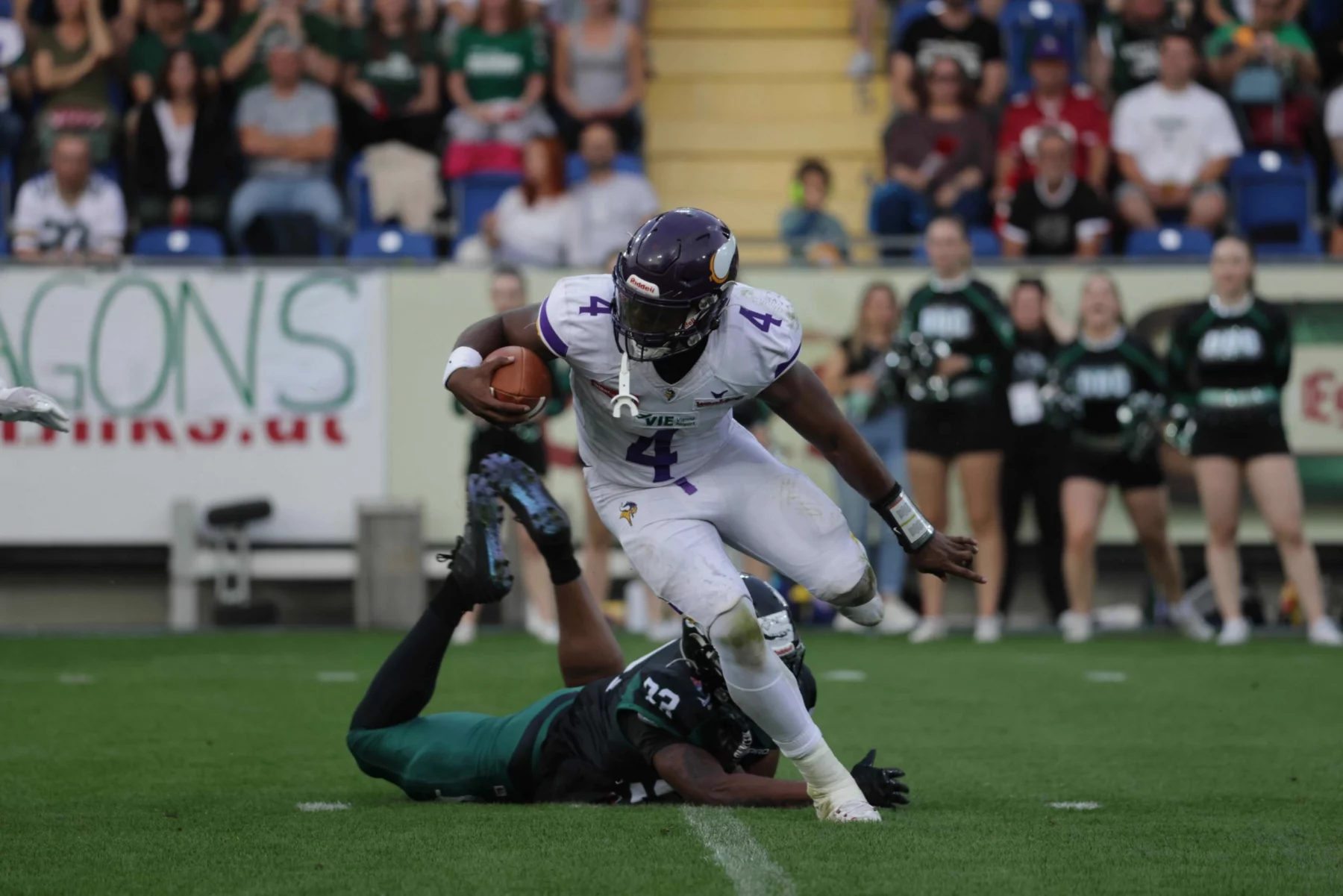 Ein Footballspieler in einem weißen Vikings-Trikot mit der Nummer 4 läuft mit dem Ball und weicht einem Tackle eines Dragons-Spielers in Grün aus, der auf dem Rasen liegt. Cheerleader und eine Menschenmenge schauen im Stadionhintergrund zu. Diese Beschreibung wurde mit der FootballR KI automatisch generiert.