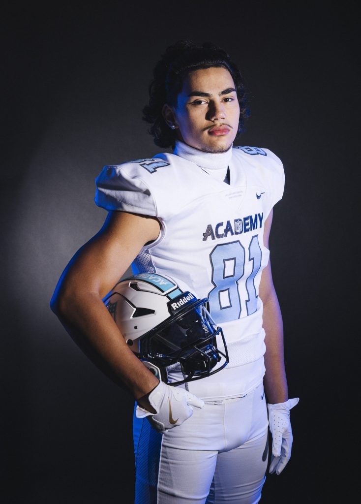 Luca Wolf - Ein junger männlicher Footballspieler steht selbstbewusst vor einem dunklen Hintergrund und trägt eine weiße NFL-Academy-Uniform mit der Aufschrift "ACADEMY" und der Nummer 81 auf seinem Trikot. Er hält einen weißen Helm an seiner Seite und blickt in die Kamera, die mit kühlem blauen Akzentlicht beleuchtet wird. Diese Beschreibung wurde mit der FootballR KI automatisch generiert.