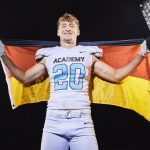 Moritz Stempel - Ein lächelnder NFL Academy Footballspieler in einer weißen Uniform mit der Aufschrift "ACADEMY" und der Nummer 20 hält eine deutsche Flagge hinter sich. Er steht zwischen zwei hellen Studiolichtern und feiert Absolventen auf dem Weg zu US-Colleges vor einem dunklen Hintergrund. Diese Beschreibung wurde mit der FootballR KI automatisch generiert.