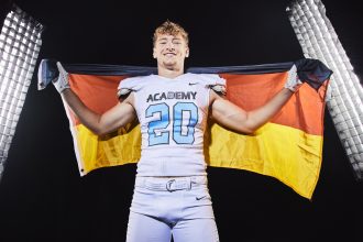 Moritz Stempel - Ein lächelnder NFL Academy Footballspieler in einer weißen Uniform mit der Aufschrift "ACADEMY" und der Nummer 20 hält eine deutsche Flagge hinter sich. Er steht zwischen zwei hellen Studiolichtern und feiert Absolventen auf dem Weg zu US-Colleges vor einem dunklen Hintergrund. Diese Beschreibung wurde mit der FootballR KI automatisch generiert.