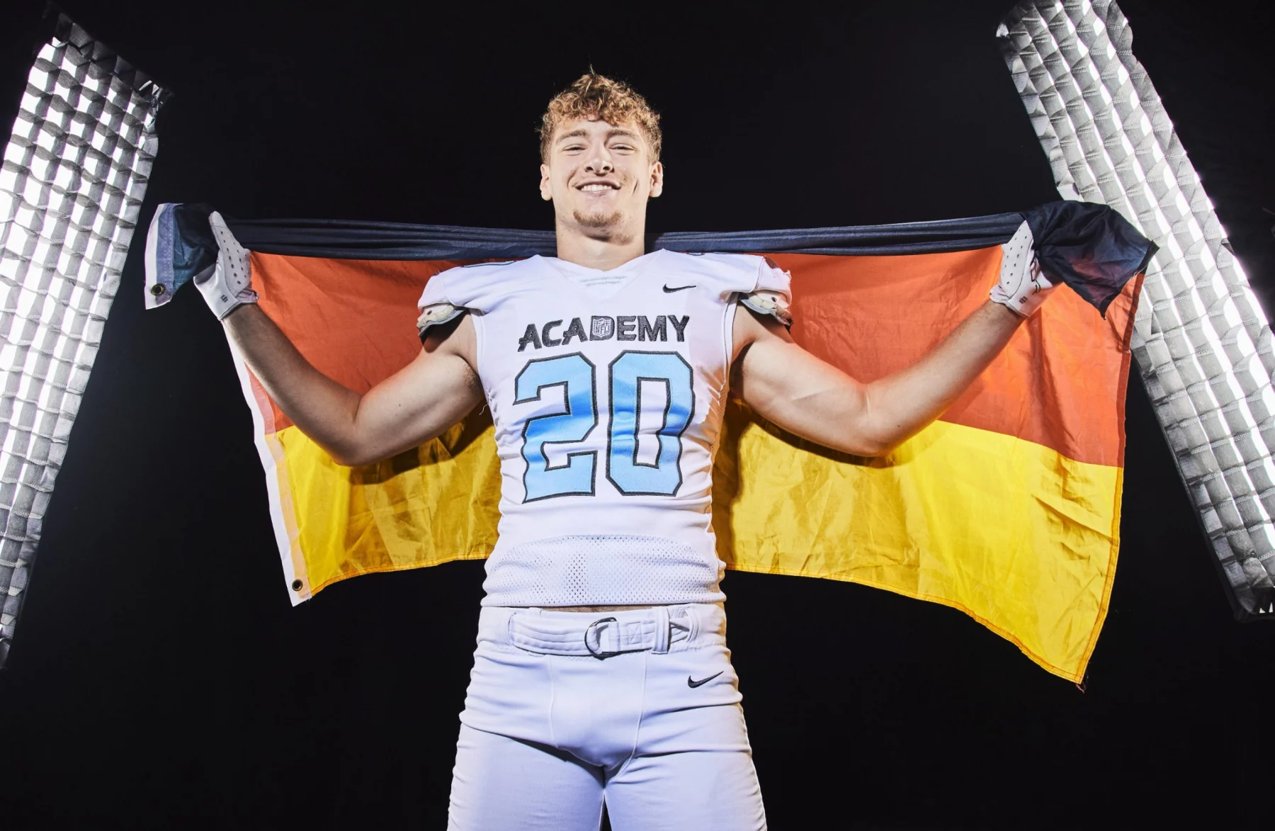 Moritz Stempel - Ein lächelnder NFL Academy Footballspieler in einer weißen Uniform mit der Aufschrift "ACADEMY" und der Nummer 20 hält eine deutsche Flagge hinter sich. Er steht zwischen zwei hellen Studiolichtern und feiert Absolventen auf dem Weg zu US-Colleges vor einem dunklen Hintergrund. Diese Beschreibung wurde mit der FootballR KI automatisch generiert.