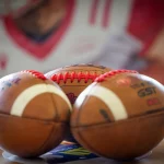 Drei amerikanische Footbälle liegen dicht nebeneinander auf einem Tisch, zwei vor und einer hinter ihnen. Im unscharfen Hintergrund ist ein großes Bild eines Footballspielers zu sehen, das auf heimische Ligen für das Football-Jahr 2026 hinweist. Diese Beschreibung wurde mit der FootballR KI automatisch generiert.