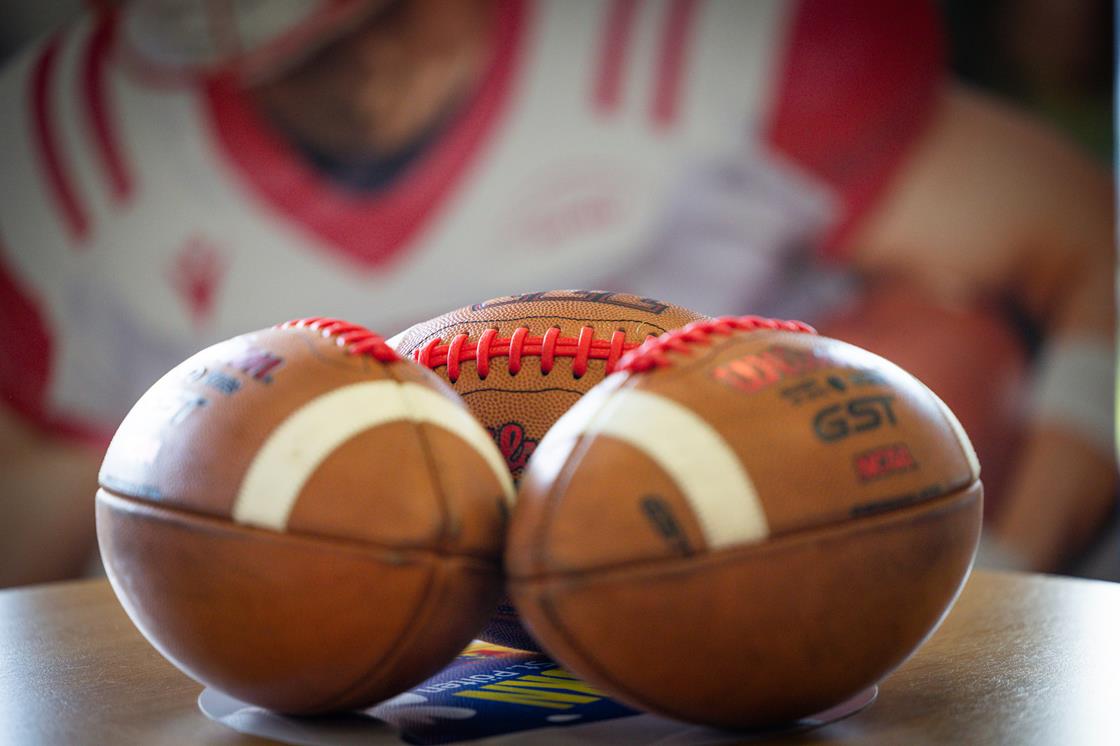 Drei amerikanische Footbälle liegen dicht nebeneinander auf einem Tisch, zwei vor und einer hinter ihnen. Im unscharfen Hintergrund ist ein großes Bild eines Footballspielers zu sehen, das auf heimische Ligen für das Football-Jahr 2026 hinweist. Diese Beschreibung wurde mit der FootballR KI automatisch generiert.