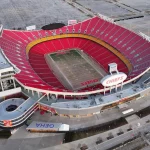 Luftaufnahme des leeren GEHA Field im Arrowhead Stadium, der Heimat der Kansas City Chiefs. Leuchtend rote Sitze umgeben das grüne Football-Feld in Kansas, mit "CHIEFS" in den Endzonen und leeren Parkplätzen rund um das Stadion. Diese Beschreibung wurde mit der FootballR KI automatisch generiert.