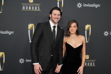 Josh Allen, in schwarzem Anzug und Krawatte, steht lächelnd neben Hailee Steinfeld in einem trägerlosen schwarzen Kleid und einer Diamantenkette auf dem roten Teppich einer NFL Honors Veranstaltung. Sie halten Händchen und posieren für Fotos vor der NFL-Kulisse. Diese Beschreibung wurde mit der FootballR KI automatisch generiert.
