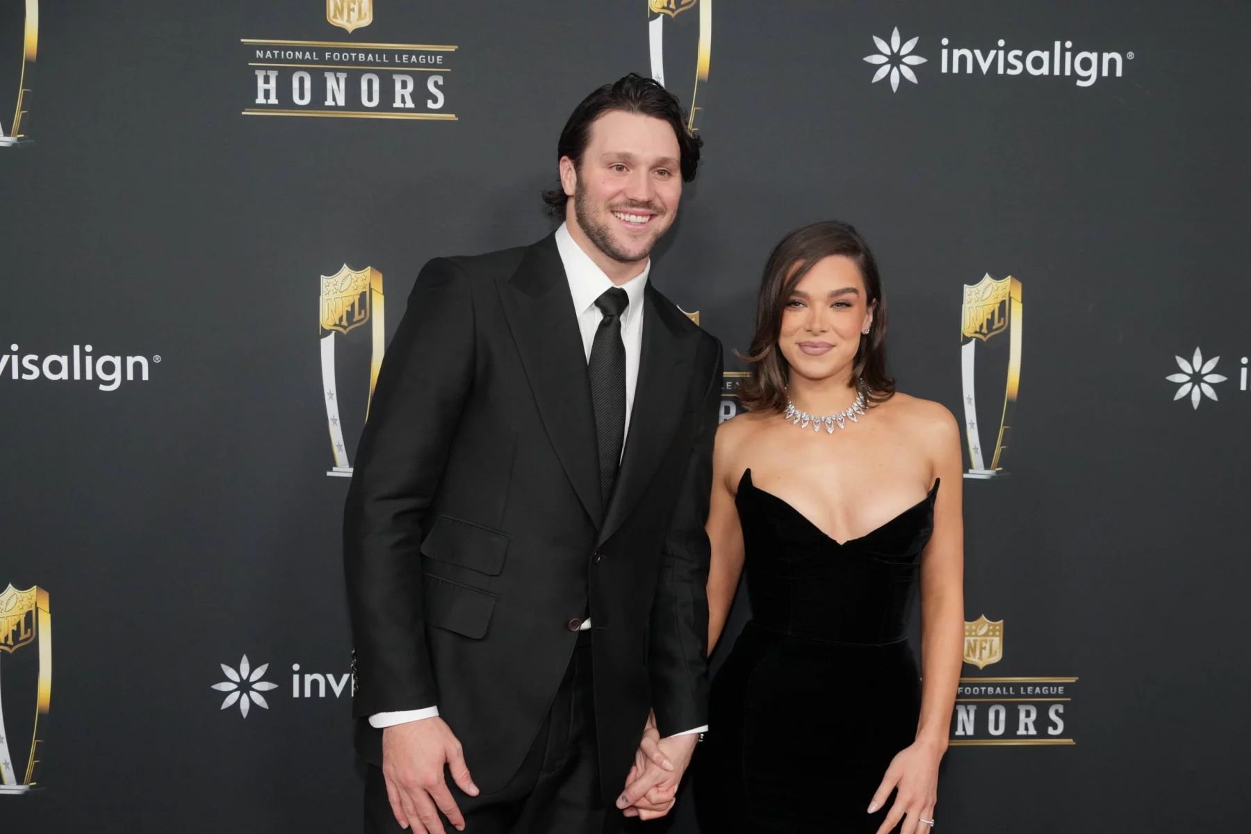 Josh Allen, in schwarzem Anzug und Krawatte, steht lächelnd neben Hailee Steinfeld in einem trägerlosen schwarzen Kleid und einer Diamantenkette auf dem roten Teppich einer NFL Honors Veranstaltung. Sie halten Händchen und posieren für Fotos vor der NFL-Kulisse. Diese Beschreibung wurde mit der FootballR KI automatisch generiert.