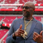 Jerry Rice - Ein Mann mit Sonnenbrille, blauem Blazer und mehreren Goldketten mit großen Anhängern steht in einem Stadion mit roten Sitzen bei den Pro Bowl Games 2026. Er lächelt und zeigt einen doppelten Daumen nach oben, während im Hintergrund unscharfe Zuschauer und helle Beleuchtung zu sehen sind. Diese Beschreibung wurde mit der FootballR KI automatisch generiert.