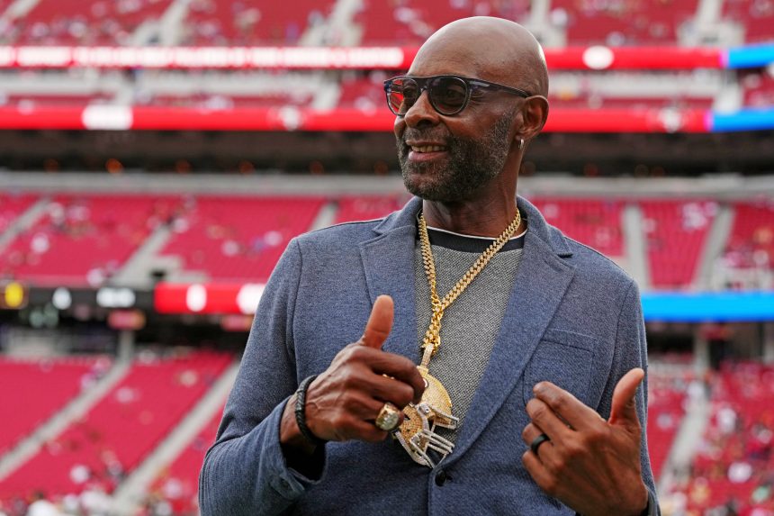 Jerry Rice - Ein Mann mit Sonnenbrille, blauem Blazer und mehreren Goldketten mit großen Anhängern steht in einem Stadion mit roten Sitzen bei den Pro Bowl Games 2026. Er lächelt und zeigt einen doppelten Daumen nach oben, während im Hintergrund unscharfe Zuschauer und helle Beleuchtung zu sehen sind. Diese Beschreibung wurde mit der FootballR KI automatisch generiert.