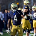 Jeremiyah Love von Notre Dame, in einem marineblauen Trikot mit der Nummer 4 und einer goldenen Hose, steht auf dem Feld und gestikuliert mit der linken Hand. Ein glänzender goldener Helm krönt seinen Look, während im unscharfen Hintergrund Trainer und ein Schiedsrichter zu sehen sind - ein Kandidat für den NFL-Draft 2026. Diese Beschreibung wurde mit der FootballR KI automatisch generiert.