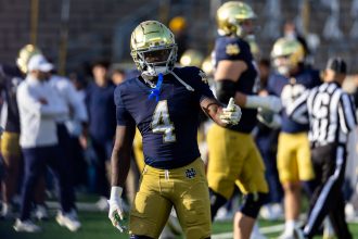 Jeremiyah Love von Notre Dame, in einem marineblauen Trikot mit der Nummer 4 und einer goldenen Hose, steht auf dem Feld und gestikuliert mit der linken Hand. Ein glänzender goldener Helm krönt seinen Look, während im unscharfen Hintergrund Trainer und ein Schiedsrichter zu sehen sind - ein Kandidat für den NFL-Draft 2026. Diese Beschreibung wurde mit der FootballR KI automatisch generiert.