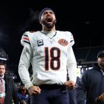 Caleb Williams - NFL Playoff Picture - Ein Footballspieler im Trikot der Chicago Bears mit der Nummer 18 schreit nachts mit geballten Fäusten leidenschaftlich auf dem Spielfeld. Er trägt ein Kapitänsabzeichen und ein Stirnband und heizt das Rennen um die NFL-Playoff-Bilder inmitten von jubelnden Fans und hellen Stadionlichtern an. Diese Beschreibung wurde mit der FootballR KI automatisch generiert.