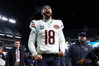 Caleb Williams - NFL Playoff Picture - Ein Footballspieler im Trikot der Chicago Bears mit der Nummer 18 schreit nachts mit geballten Fäusten leidenschaftlich auf dem Spielfeld. Er trägt ein Kapitänsabzeichen und ein Stirnband und heizt das Rennen um die NFL-Playoff-Bilder inmitten von jubelnden Fans und hellen Stadionlichtern an. Diese Beschreibung wurde mit der FootballR KI automatisch generiert.