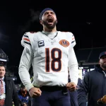 Caleb Williams - NFL Playoff Picture - Ein Footballspieler im Trikot der Chicago Bears mit der Nummer 18 schreit nachts mit geballten Fäusten leidenschaftlich auf dem Spielfeld. Er trägt ein Kapitänsabzeichen und ein Stirnband und heizt das Rennen um die NFL-Playoff-Bilder inmitten von jubelnden Fans und hellen Stadionlichtern an. Diese Beschreibung wurde mit der FootballR KI automatisch generiert.