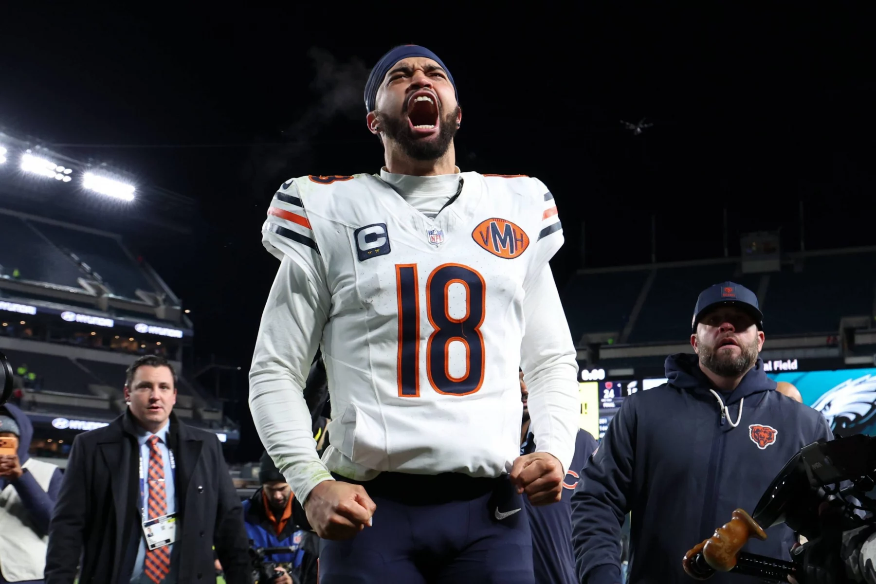 Caleb Williams - NFL Playoff Picture - Ein Footballspieler im Trikot der Chicago Bears mit der Nummer 18 schreit nachts mit geballten Fäusten leidenschaftlich auf dem Spielfeld. Er trägt ein Kapitänsabzeichen und ein Stirnband und heizt das Rennen um die NFL-Playoff-Bilder inmitten von jubelnden Fans und hellen Stadionlichtern an. Diese Beschreibung wurde mit der FootballR KI automatisch generiert.
