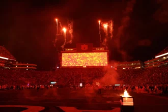 Targeting Strafe: Ein großes Stadion voller Fans bei Nacht. Der Himmel leuchtet rot vom Feuerwerk über einer riesigen hellen Anzeigetafel mit der Aufschrift "Auburn Tigers". Die Stadionbeleuchtung und die Flammen in der Nähe des Spielfelds tragen zu der dramatischen College-Football-Szene bei, und die Tribünen sind mit silhouettierten Zuschauern gefüllt. Diese Beschreibung wurde mit der FootballR KI automatisch generiert.