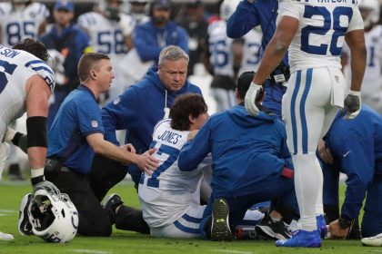 Football-Trainer und Mannschaftskameraden versammeln sich um den Colts-Spieler Daniel Jones, der nach einem offensichtlichen Achillessehnenriss auf dem Spielfeld sitzt. Auf dem Trikot des Spielers steht "JONES 7". Medizinisches Personal kümmert sich um ihn, während besorgte Teamkollegen zuschauen. Diese Beschreibung wurde mit der FootballR KI automatisch generiert.