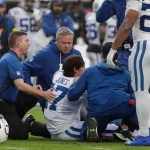 Football-Trainer und Mannschaftskameraden versammeln sich um den Colts-Spieler Daniel Jones, der nach einem offensichtlichen Achillessehnenriss auf dem Spielfeld sitzt. Auf dem Trikot des Spielers steht "JONES 7". Medizinisches Personal kümmert sich um ihn, während besorgte Teamkollegen zuschauen. Diese Beschreibung wurde mit der FootballR KI automatisch generiert.