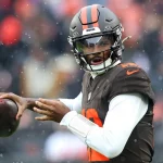 Ein Football-Spieler der Browns in Uniform und Helm bereitet sich darauf vor, bei nassem, regnerischem Wetter einen Football zu werfen. Im Hintergrund sind unscharfe Fans zu sehen, die die Intensität und Entschlossenheit der Zukunft der Franchise widerspiegeln. Diese Beschreibung wurde mit der FootballR KI automatisch generiert.