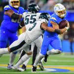 Justin Herbert - Ein Football-Spieler in einem blauen Chargers-Trikot läuft mit dem Ball, während er von einem Eagles-Spieler in einem weiß-grünen Trikot um die Hüfte angegriffen wird. Im Hintergrund sind andere Spieler und unscharfe Zuschauer auf einem hell erleuchteten Football-Feld zu sehen. Diese Beschreibung wurde mit der FootballR KI automatisch generiert.