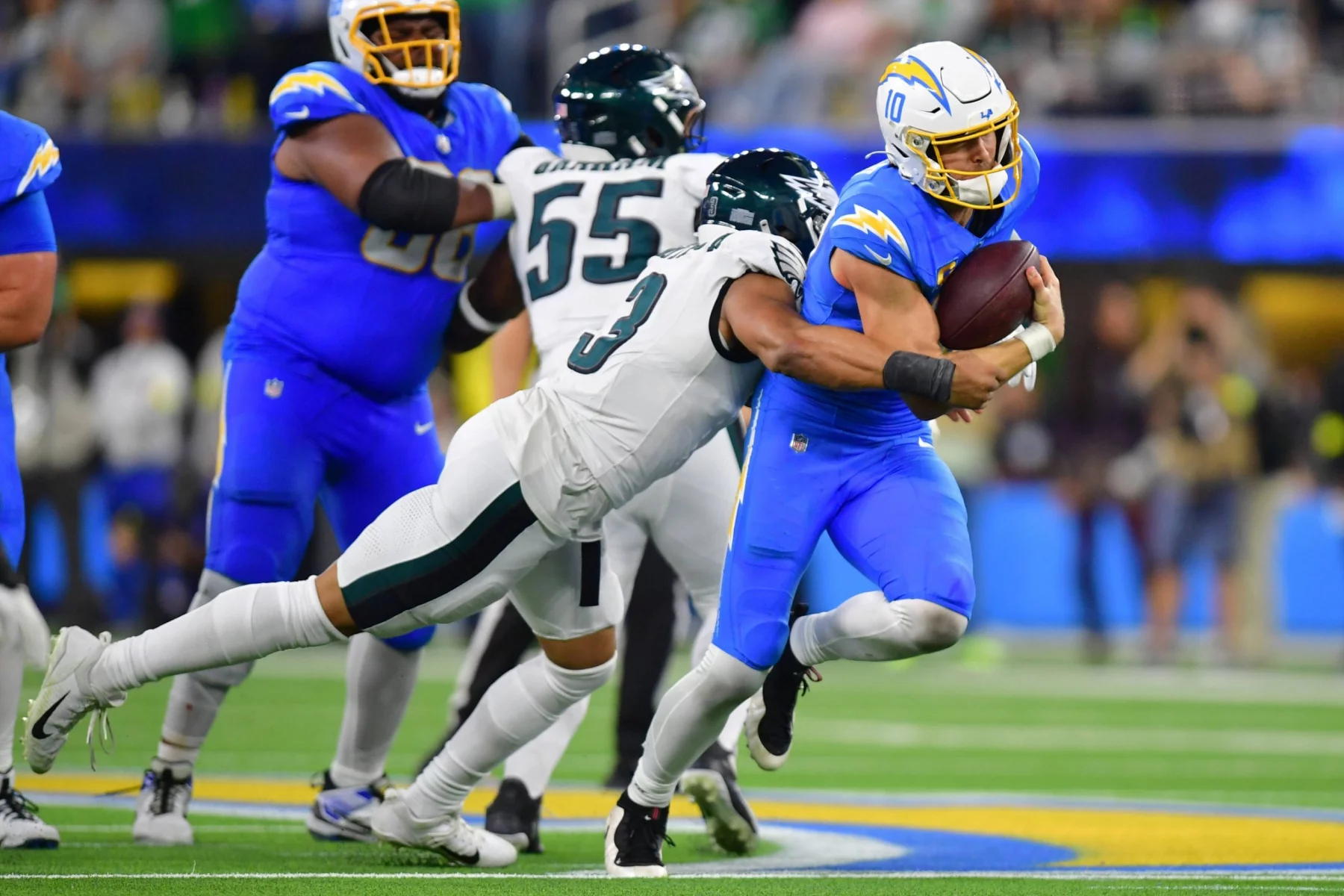 Justin Herbert - Ein Football-Spieler in einem blauen Chargers-Trikot läuft mit dem Ball, während er von einem Eagles-Spieler in einem weiß-grünen Trikot um die Hüfte angegriffen wird. Im Hintergrund sind andere Spieler und unscharfe Zuschauer auf einem hell erleuchteten Football-Feld zu sehen. Diese Beschreibung wurde mit der FootballR KI automatisch generiert.