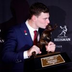 Fernando Mendoza, der eine rote Krawatte und eine Anstecknadel der Indiana University trägt, küsst die Heisman Trophy 2025 und hält sie mit beiden Händen. Im Hintergrund sind wiederholte Heisman-Logos zu sehen, und die Plakette der Trophäe ist deutlich zu erkennen. Diese Beschreibung wurde mit der FootballR KI automatisch generiert.