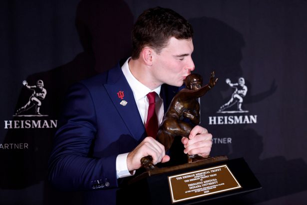 Fernando Mendoza, der eine rote Krawatte und eine Anstecknadel der Indiana University trägt, küsst die Heisman Trophy 2025 und hält sie mit beiden Händen. Im Hintergrund sind wiederholte Heisman-Logos zu sehen, und die Plakette der Trophäe ist deutlich zu erkennen. Diese Beschreibung wurde mit der FootballR KI automatisch generiert.