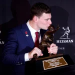 Fernando Mendoza, der eine rote Krawatte und eine Anstecknadel der Indiana University trägt, küsst die Heisman Trophy 2025 und hält sie mit beiden Händen. Im Hintergrund sind wiederholte Heisman-Logos zu sehen, und die Plakette der Trophäe ist deutlich zu erkennen. Diese Beschreibung wurde mit der FootballR KI automatisch generiert.