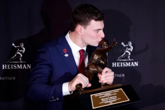 Fernando Mendoza, der eine rote Krawatte und eine Anstecknadel der Indiana University trägt, küsst die Heisman Trophy 2025 und hält sie mit beiden Händen. Im Hintergrund sind wiederholte Heisman-Logos zu sehen, und die Plakette der Trophäe ist deutlich zu erkennen. Diese Beschreibung wurde mit der FootballR KI automatisch generiert.