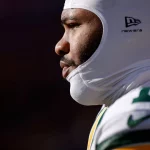 Micah Parsons - Nahaufnahme, Seitenprofil eines Packers-Footballspielers, der einen weißen, gepolsterten Helm und eine grün-gelbe Uniform trägt. Der Spieler blickt konzentriert nach links, wobei helles Sonnenlicht sein Gesicht vor einem dunklen, unscharfen Hintergrund hervorhebt. Diese Beschreibung wurde mit der FootballR KI automatisch generiert.