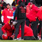 Star-QB Patrick Mahomes verletzt - Mitarbeiter und Trainer der Kansas City Chiefs in roten Jacken umringen ihren Star-QB Patrick Mahomes, der verletzt auf dem Spielfeld liegt. Ein Mitarbeiter kniet nieder, um nach ihm zu sehen, während die Zuschauer besorgt auf einen möglichen Kreuzbandriss blicken. Diese Beschreibung wurde mit der FootballR KI automatisch generiert.