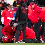 Star-QB Patrick Mahomes verletzt - Mitarbeiter und Trainer der Kansas City Chiefs in roten Jacken umringen ihren Star-QB Patrick Mahomes, der verletzt auf dem Spielfeld liegt. Ein Mitarbeiter kniet nieder, um nach ihm zu sehen, während die Zuschauer besorgt auf einen möglichen Kreuzbandriss blicken. Diese Beschreibung wurde mit der FootballR KI automatisch generiert.