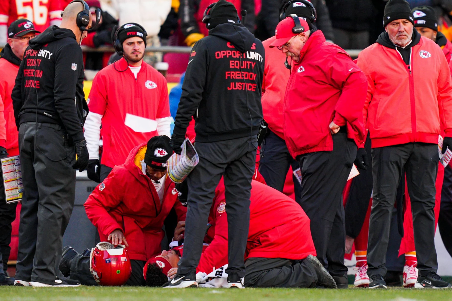 Star-QB Patrick Mahomes verletzt - Mitarbeiter und Trainer der Kansas City Chiefs in roten Jacken umringen ihren Star-QB Patrick Mahomes, der verletzt auf dem Spielfeld liegt. Ein Mitarbeiter kniet nieder, um nach ihm zu sehen, während die Zuschauer besorgt auf einen möglichen Kreuzbandriss blicken. Diese Beschreibung wurde mit der FootballR KI automatisch generiert.
