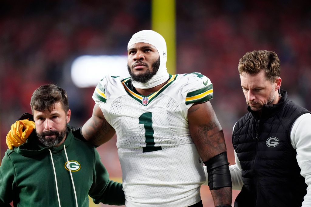 Micah Parsons - Ein Football-Spieler der Green Bay Packers in einem weißen Trikot mit der Nummer 1, der einen Verband am Kopf trägt, wird von zwei Mitarbeitern gestützt, als er das Spielfeld verlässt - möglicherweise durch eine Verletzung wie ein Kreuzbandriss außer Gefecht gesetzt - inmitten verschwommener roter Stadionlichter bei Nacht. Diese Beschreibung wurde mit der FootballR KI automatisch generiert.