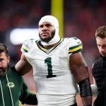 Micah Parsons - Ein Football-Spieler der Green Bay Packers in einem weißen Trikot mit der Nummer 1, der einen Verband am Kopf trägt, wird von zwei Mitarbeitern gestützt, als er das Spielfeld verlässt - möglicherweise durch eine Verletzung wie ein Kreuzbandriss außer Gefecht gesetzt - inmitten verschwommener roter Stadionlichter bei Nacht. Diese Beschreibung wurde mit der FootballR KI automatisch generiert.