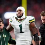 Micah Parsons - Ein Football-Spieler der Green Bay Packers in einem weißen Trikot mit der Nummer 1, der einen Verband am Kopf trägt, wird von zwei Mitarbeitern gestützt, als er das Spielfeld verlässt - möglicherweise durch eine Verletzung wie ein Kreuzbandriss außer Gefecht gesetzt - inmitten verschwommener roter Stadionlichter bei Nacht. Diese Beschreibung wurde mit der FootballR KI automatisch generiert.