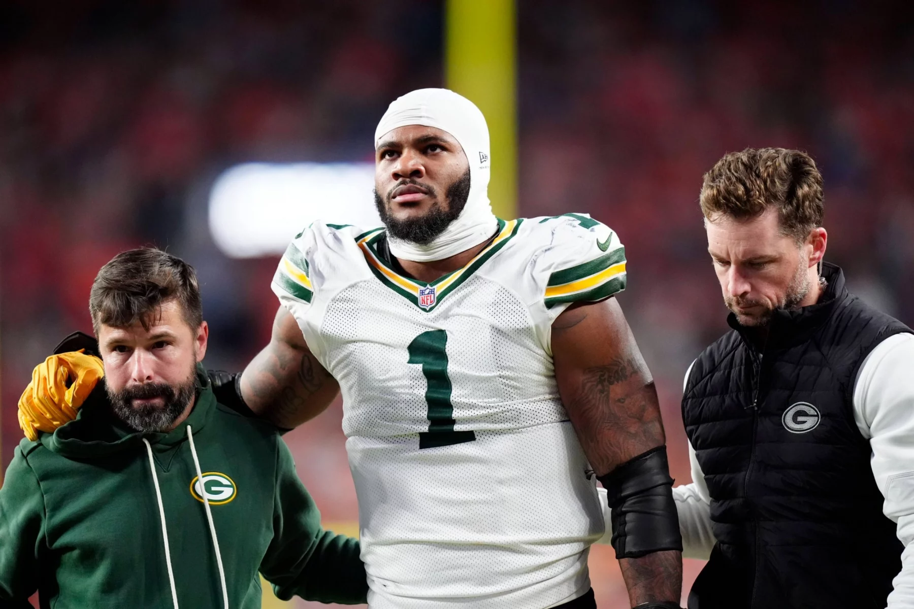 Micah Parsons - Ein Football-Spieler der Green Bay Packers in einem weißen Trikot mit der Nummer 1, der einen Verband am Kopf trägt, wird von zwei Mitarbeitern gestützt, als er das Spielfeld verlässt - möglicherweise durch eine Verletzung wie ein Kreuzbandriss außer Gefecht gesetzt - inmitten verschwommener roter Stadionlichter bei Nacht. Diese Beschreibung wurde mit der FootballR KI automatisch generiert.