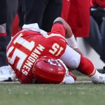 Ein Football-Spieler in einem roten Kansas City Chiefs-Trikot mit der Nummer 15 liegt mit dem Gesicht nach unten, seinen Helm umklammernd, auf dem Feld, umgeben von Spielern und Personal an der Seitenlinie, was auf einen möglichen Kreuzbandriss und eine ungewisse Rückkehr zum Spiel hindeutet. Diese Beschreibung wurde mit der FootballR KI automatisch generiert.