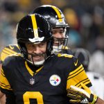 Aaron Rodgers - Ein lächelnder Footballspieler in einer schwarz-goldenen Pittsburgh Steelers-Uniform mit Helm und der Nummer 8 steht auf dem Spielfeld. Ein anderer Mannschaftskamerad ist teilweise hinter ihm zu sehen, während im Hintergrund unscharfe Zuschauer zu sehen sind, die an einen klassischen Steelers-Moment erinnern. Diese Beschreibung wurde mit der FootballR KI automatisch generiert.