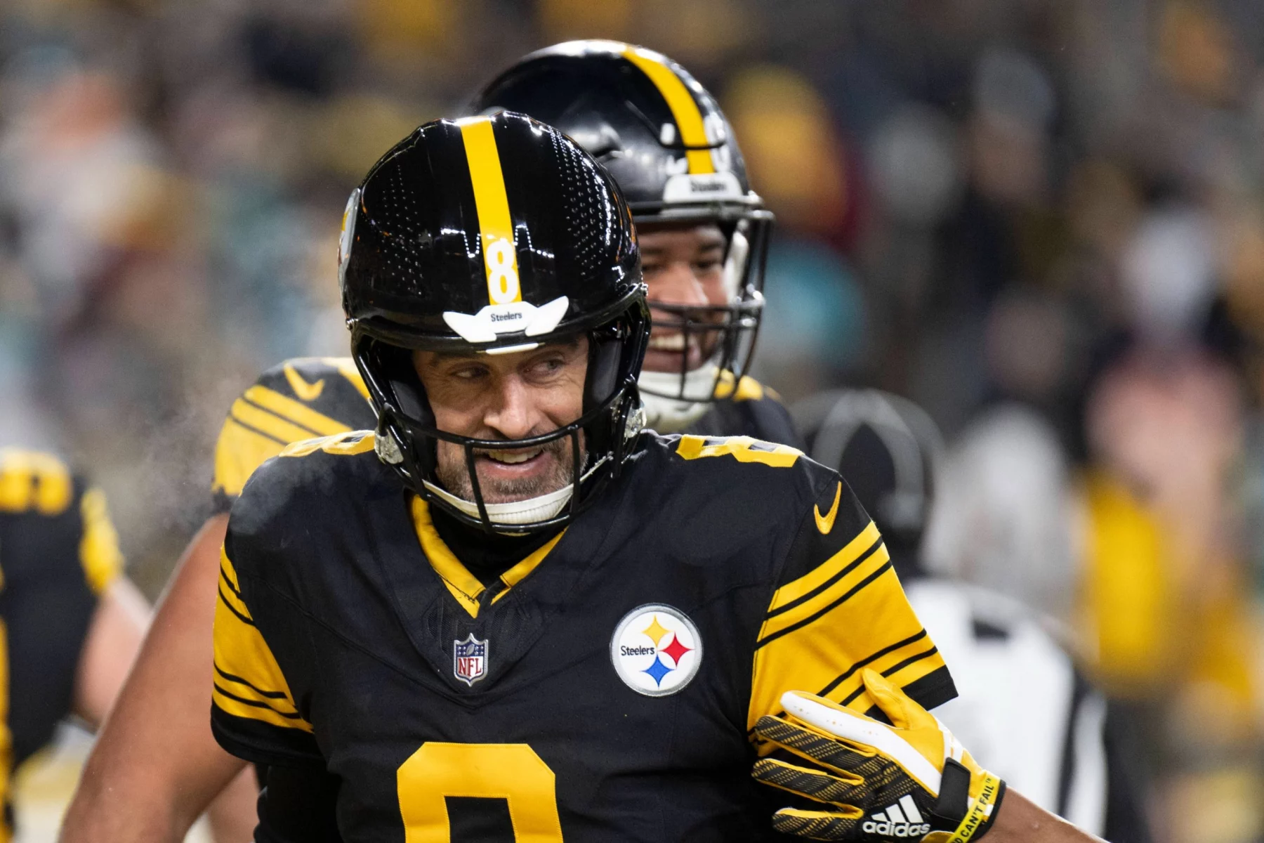 Aaron Rodgers - Ein lächelnder Footballspieler in einer schwarz-goldenen Pittsburgh Steelers-Uniform mit Helm und der Nummer 8 steht auf dem Spielfeld. Ein anderer Mannschaftskamerad ist teilweise hinter ihm zu sehen, während im Hintergrund unscharfe Zuschauer zu sehen sind, die an einen klassischen Steelers-Moment erinnern. Diese Beschreibung wurde mit der FootballR KI automatisch generiert.