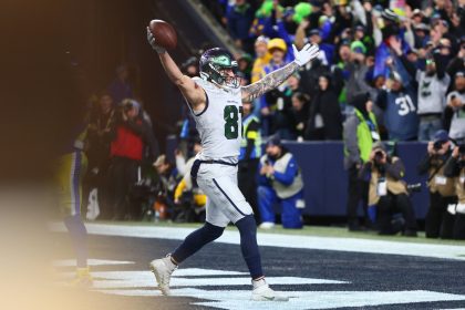 Ein Footballspieler der Seattle Seahawks in weißer und marineblauer Uniform feiert in der Endzone mit erhobenen Armen und hält einen Football. Die Menge jubelt, als das Team sich das Playoff-Ticket sichert, und fängt einen Moment des Triumphs während des Spiels ein. Diese Beschreibung wurde mit der FootballR KI automatisch generiert.
