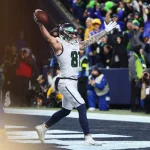 Ein Footballspieler der Seattle Seahawks in weißer und marineblauer Uniform feiert in der Endzone mit erhobenen Armen und hält einen Football. Die Menge jubelt, als das Team sich das Playoff-Ticket sichert, und fängt einen Moment des Triumphs während des Spiels ein. Diese Beschreibung wurde mit der FootballR KI automatisch generiert.