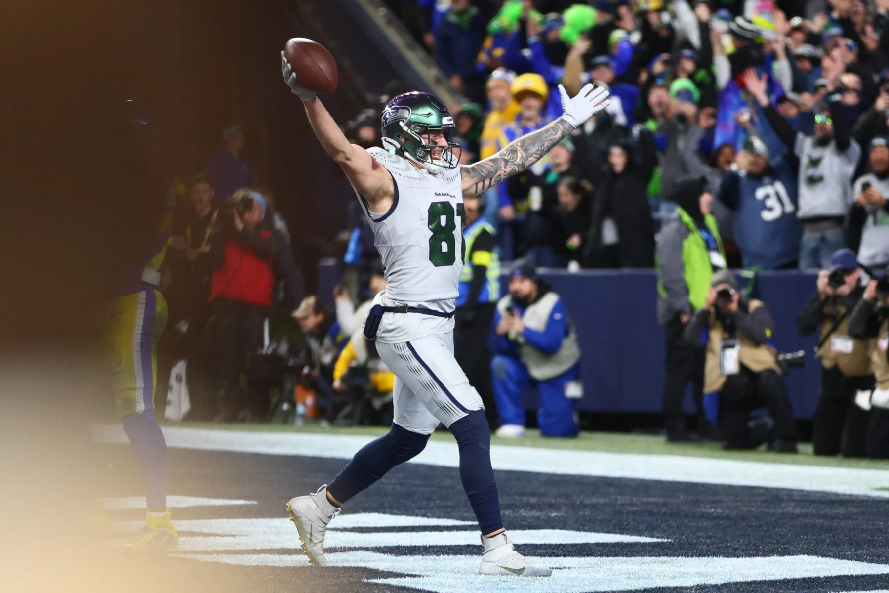Ein Footballspieler der Seattle Seahawks in weißer und marineblauer Uniform feiert in der Endzone mit erhobenen Armen und hält einen Football. Die Menge jubelt, als das Team sich das Playoff-Ticket sichert, und fängt einen Moment des Triumphs während des Spiels ein. Diese Beschreibung wurde mit der FootballR KI automatisch generiert.