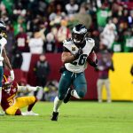 Ein NFL-Spieler in einem weiß-grünen Eagles-Trikot läuft während eines NFC East-Spiels mit dem Football. Ein anderer Eagles-Spieler hebt einen Arm, während ein Verteidiger der Washington Commanders am Boden sitzt. Fans in Jacken füllen die Tribünen im Hintergrund. Diese Beschreibung wurde mit der FootballR KI automatisch generiert.