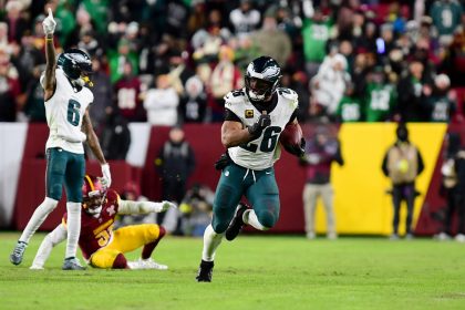 Ein NFL-Spieler in einem weiß-grünen Eagles-Trikot läuft während eines NFC East-Spiels mit dem Football. Ein anderer Eagles-Spieler hebt einen Arm, während ein Verteidiger der Washington Commanders am Boden sitzt. Fans in Jacken füllen die Tribünen im Hintergrund. Diese Beschreibung wurde mit der FootballR KI automatisch generiert.