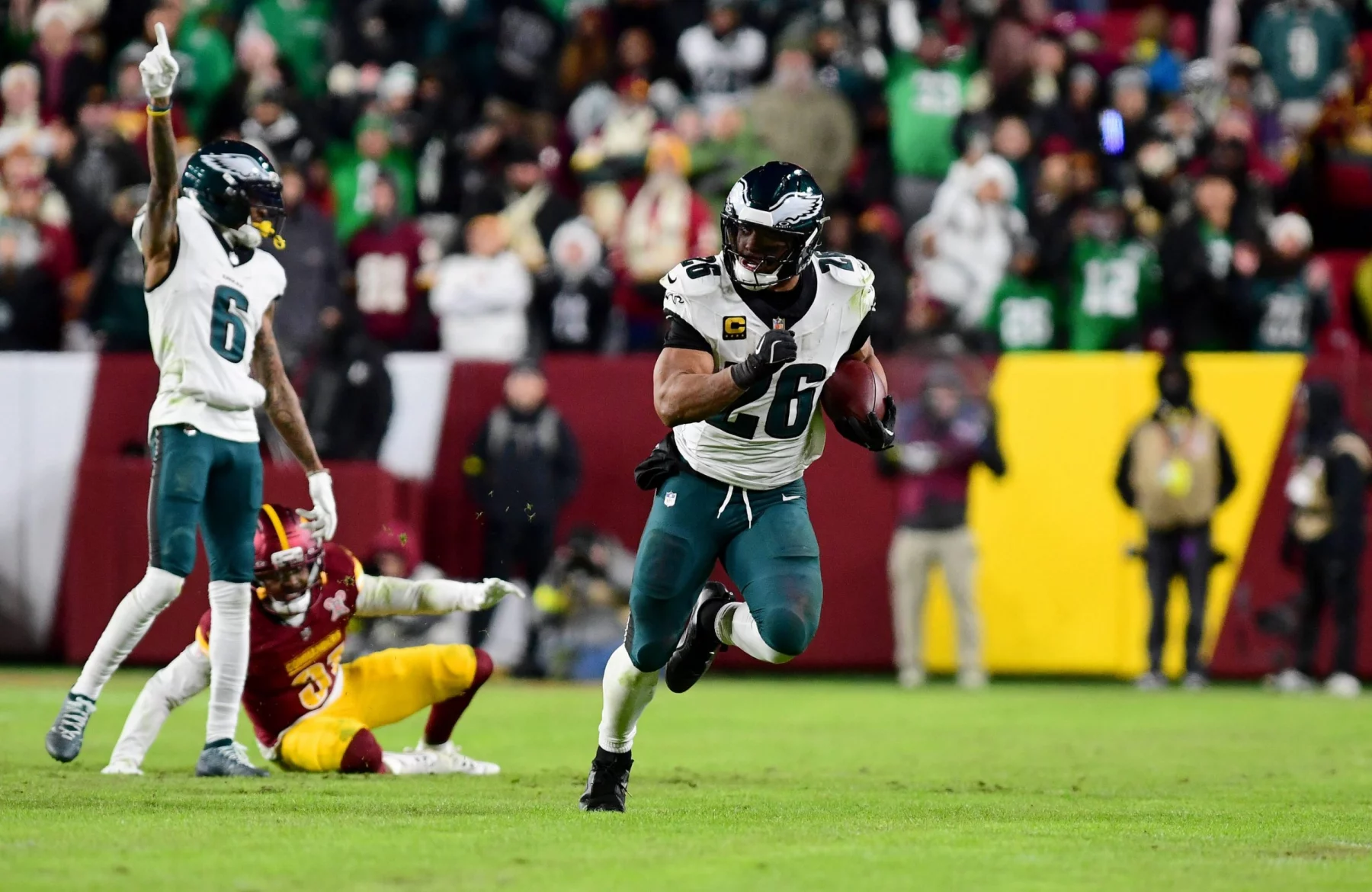Ein NFL-Spieler in einem weiß-grünen Eagles-Trikot läuft während eines NFC East-Spiels mit dem Football. Ein anderer Eagles-Spieler hebt einen Arm, während ein Verteidiger der Washington Commanders am Boden sitzt. Fans in Jacken füllen die Tribünen im Hintergrund. Diese Beschreibung wurde mit der FootballR KI automatisch generiert.