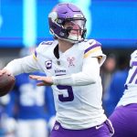Der Quarterback der Minnesota Vikings, J.J. McCarthy, in weißer und lila Uniform, bereitet sich darauf vor, während eines Spiels einen Football zu werfen. Er trägt einen lilafarbenen Helm und ein lilafarbenes Visier, neben ihm steht ein Teamkollege. Im unscharfen Hintergrund sind blau gekleidete Spieler und eine Anzeigetafel zu sehen. Diese Beschreibung wurde mit der FootballR KI automatisch generiert.