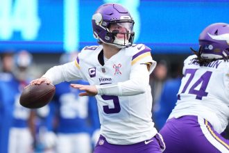 Der Quarterback der Minnesota Vikings, J.J. McCarthy, in weißer und lila Uniform, bereitet sich darauf vor, während eines Spiels einen Football zu werfen. Er trägt einen lilafarbenen Helm und ein lilafarbenes Visier, neben ihm steht ein Teamkollege. Im unscharfen Hintergrund sind blau gekleidete Spieler und eine Anzeigetafel zu sehen. Diese Beschreibung wurde mit der FootballR KI automatisch generiert.