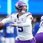 Der Quarterback der Minnesota Vikings, J.J. McCarthy, in weißer und lila Uniform, bereitet sich darauf vor, während eines Spiels einen Football zu werfen. Er trägt einen lilafarbenen Helm und ein lilafarbenes Visier, neben ihm steht ein Teamkollege. Im unscharfen Hintergrund sind blau gekleidete Spieler und eine Anzeigetafel zu sehen. Diese Beschreibung wurde mit der FootballR KI automatisch generiert.