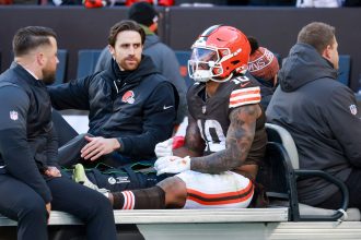 Ein Footballspieler der Browns, der Quinshon Judkins ähnelt und die Uniformnummer 10 trägt, sitzt auf einem Krankenwagen, neben ihm die Trainer. Sein linkes Bein ist gestreckt und gewickelt, was auf eine Beinverletzung während eines Spiels an der kalten Seitenlinie hinweist. Diese Beschreibung wurde mit der FootballR KI automatisch generiert.